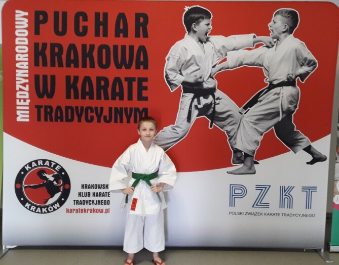 Uczeń w kimono z zielonym pasem na tle baneru Międzynarodowy Puchar Krakowa w Karate Tradycyjnym
